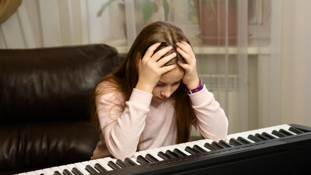 A young girl sits at a keyboard, holding her head in frustration while looking down at the keys.
