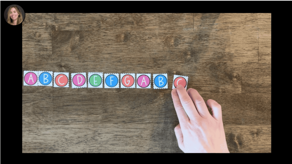 A hand reaching towards a row of colorful letter tiles displaying the letters A, B, C, D, E, F, G, A, B arranged on a wooden surface.